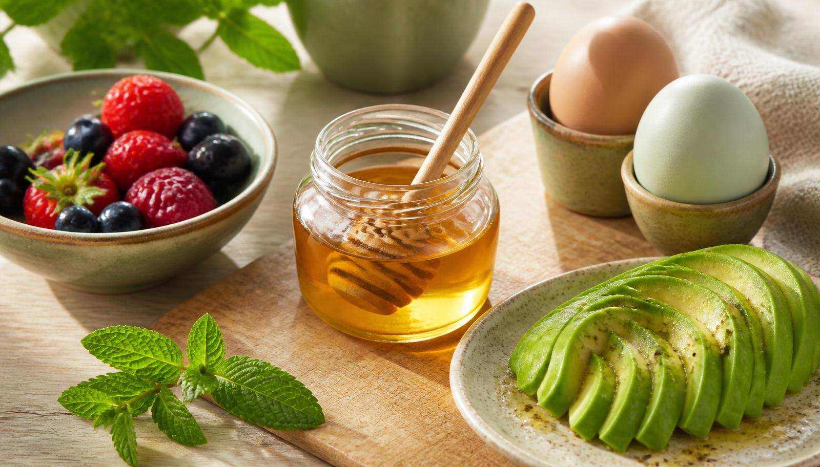 A jar of golden honey surrounded by fresh low-carb breakfast ingredients on a light wood surface