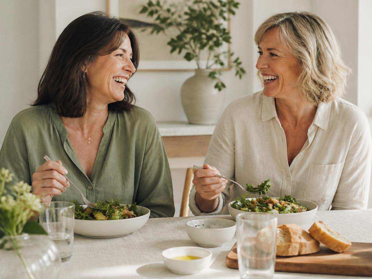 Two women in their fifties laughing together over a shared healthy lunch